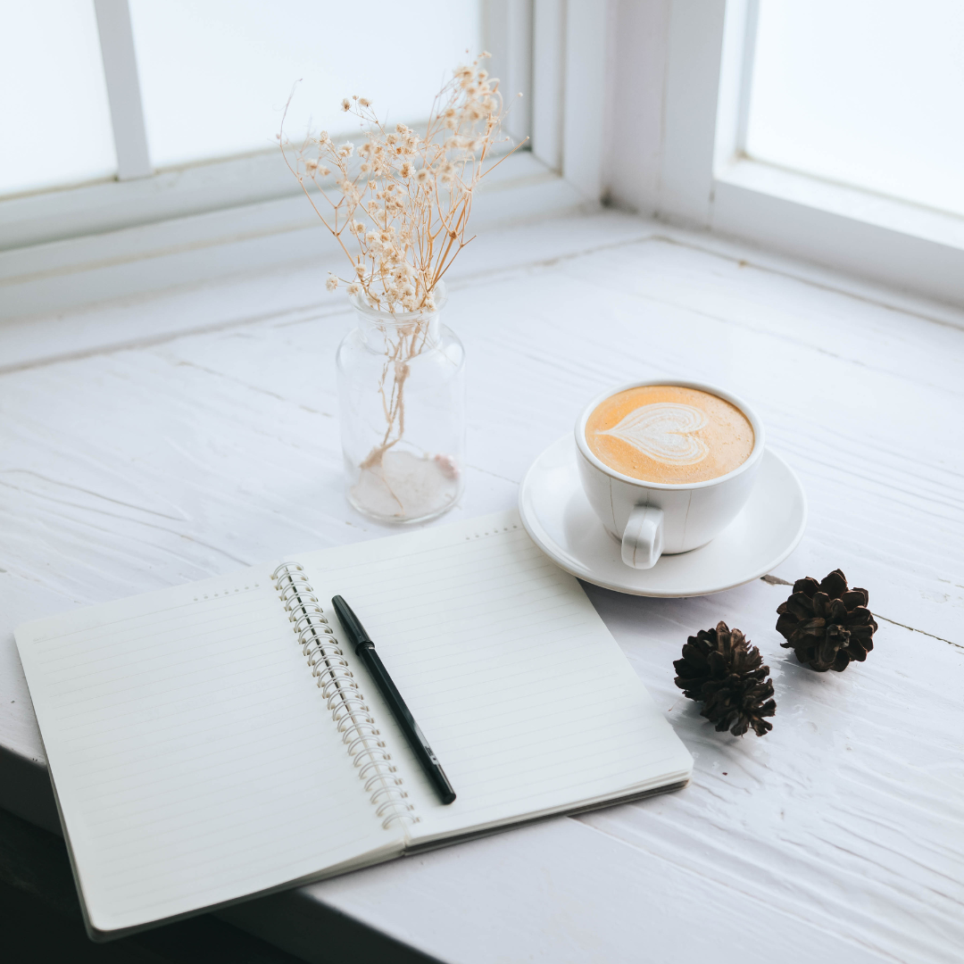 Weißer Tisch vor einem Fenster mit einem Notizbuch mit Stift, einer Tasse Kaffee, eine dekorative Pflanze und dekorative Elemente drauf.