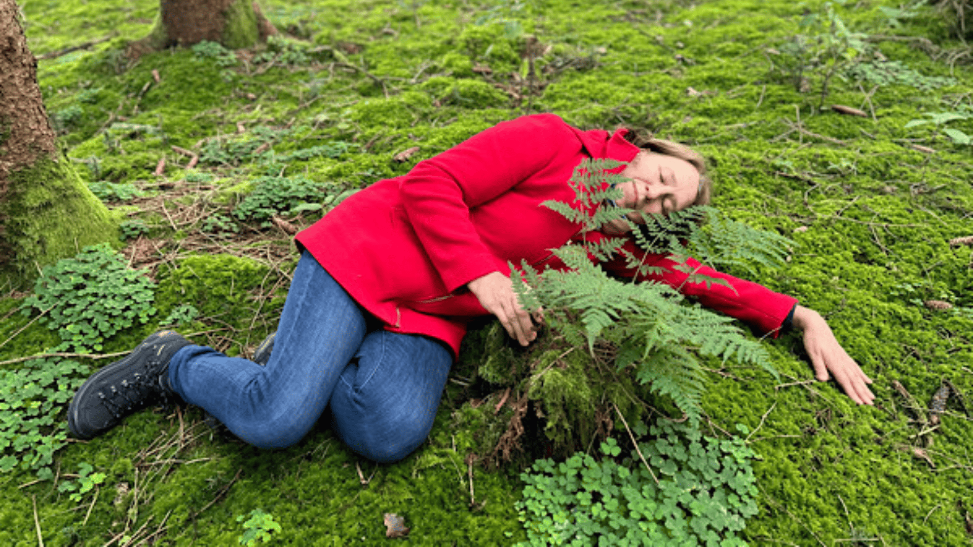 Eine Frau mit dunkelbraunen Haaren, die eine rote Jacke und Jeans trägt und im Wald auf dem Boden liegt