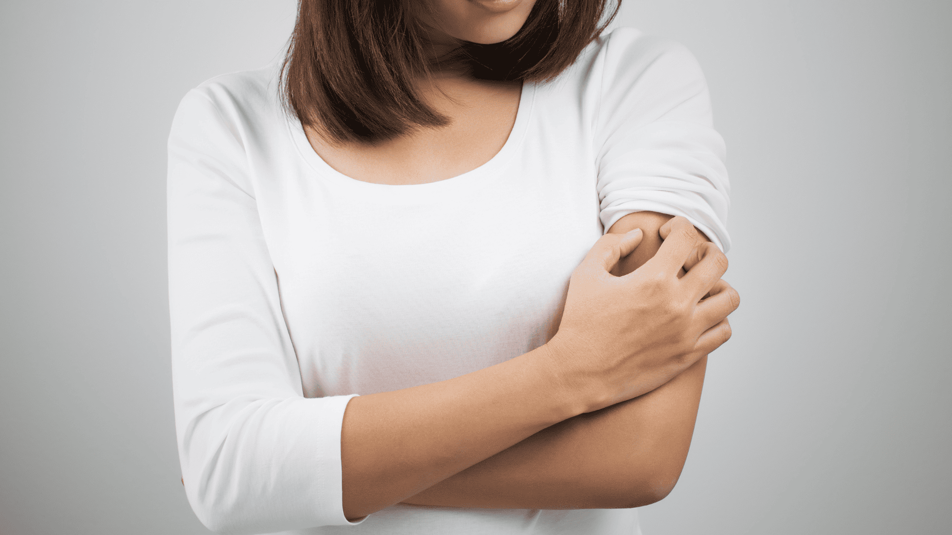 Eine Frau mit weißem T-Shirt und braunen kurzen Haaren, die sich am linken Oberarm kratzt