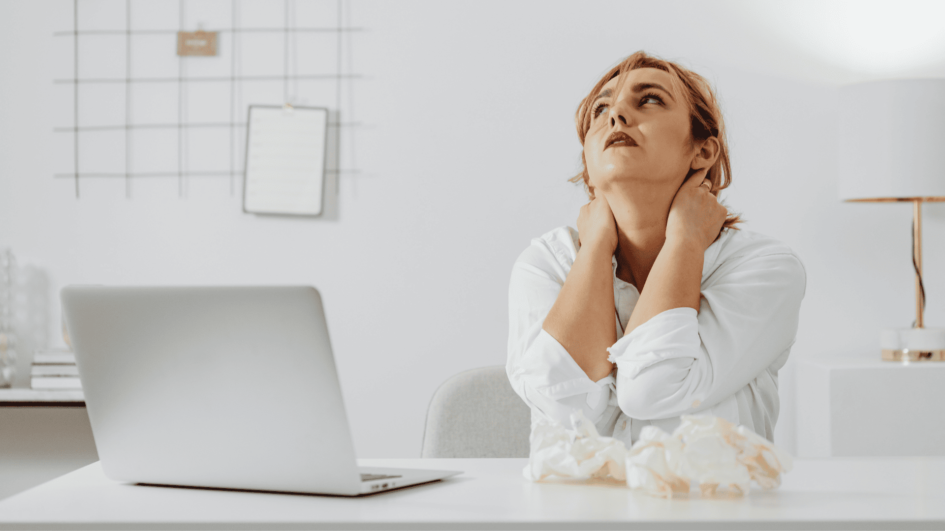 Frau, die weiß angezogen ist, vor einem weißen Schreibtisch und Laptop sitzt, nach oben sieht und aufgrund von Stress beide Hände im Nacken hält