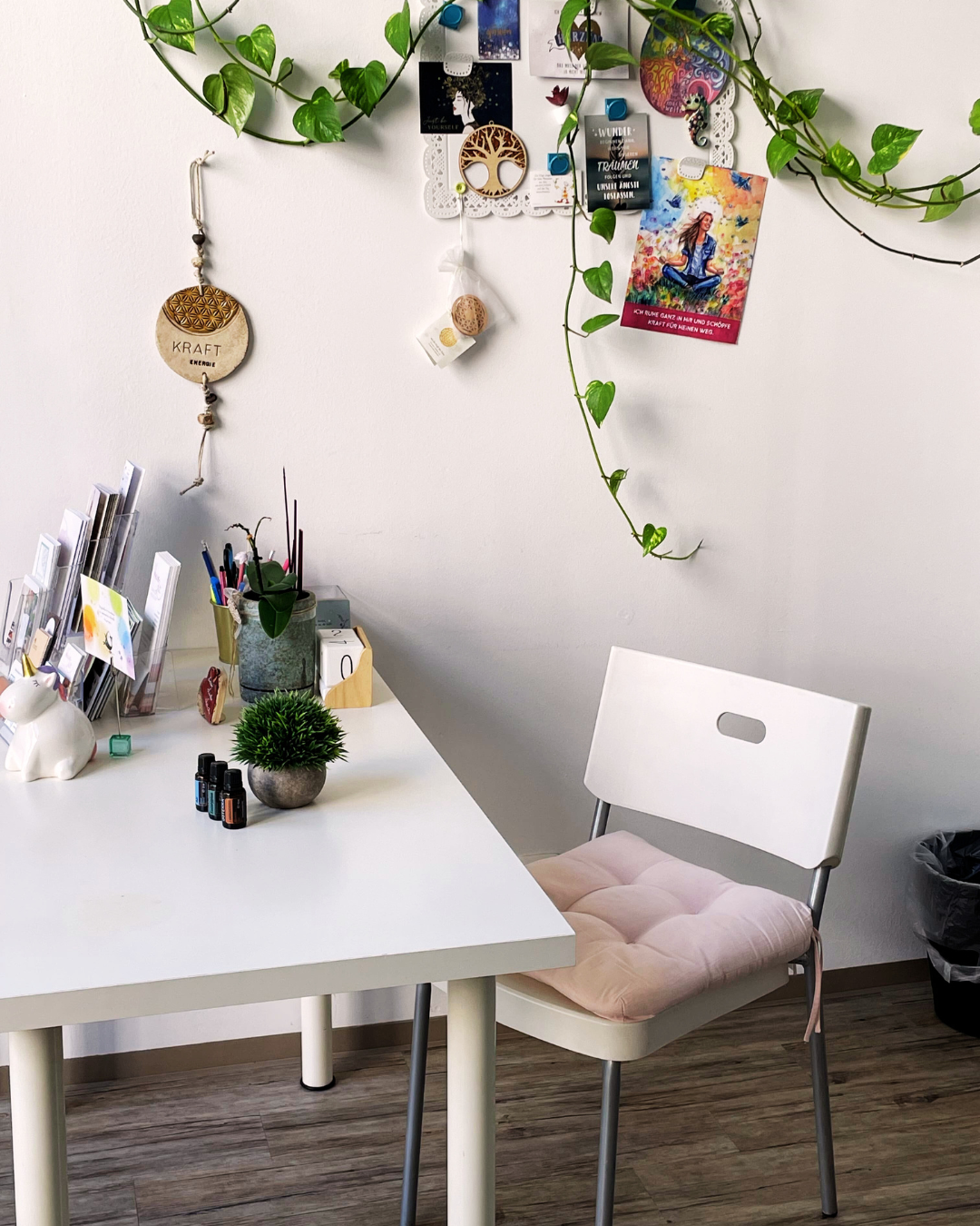 Kinesiologie Weißer Tisch mit einem weißen Stuhl, auf dem Tisch stehen ätherische Öle, eine Pflanze und ein Ständer mit diversen Werbematerialen, dahinter hängen Fotos, eine Hängepflanze und ein Symbol mit der Aufschrift Kraft - Energie
