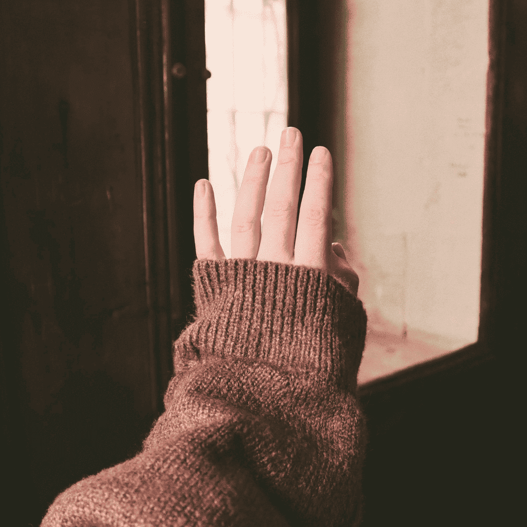 Ein Mädchenarm in einem braunen Pullover, der in Richtung eines Fensters ausgestreckt ist.