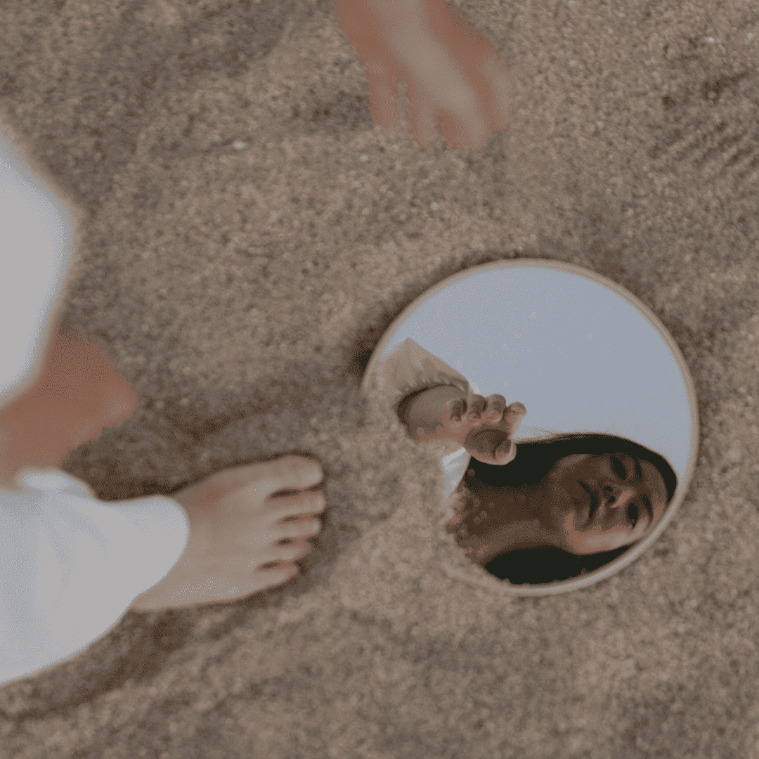 Eine Frau, die nach unten in einen Spiegel sieht, während sie an einem Strand steht und zum Spiegel hin zeigt.