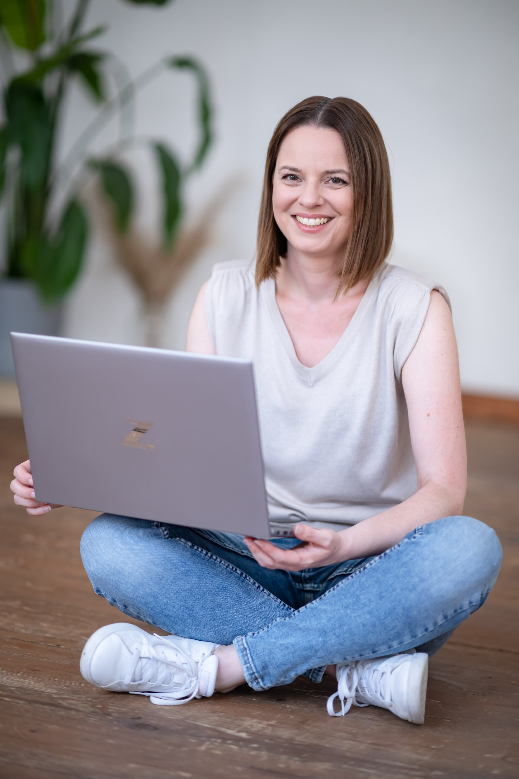 Eine Frau mit dunklen kurzen Haaren, einem beigen Shirt, Jeans, weiße Turnschuhe, die auf dem Boden sitzt und einen aufgeklappten Laptop in der Hand hält.