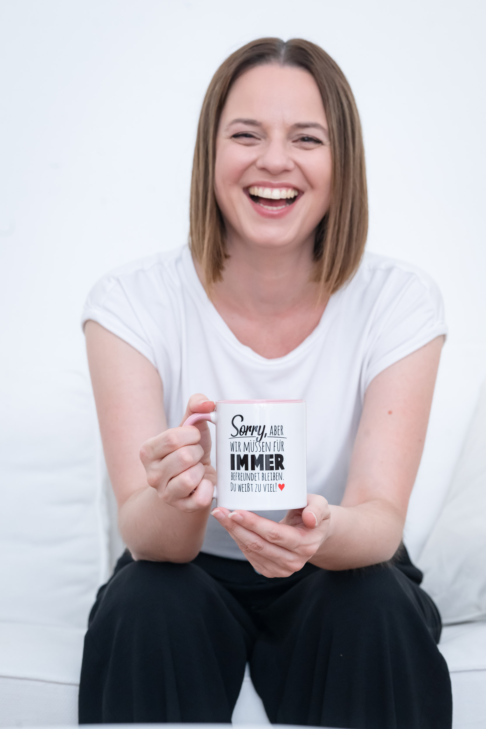 Über mich Eine Frau mit kurzen dunklen Haaren, einem weißen T-Shirt und einer schwarzen Hose, die auf einer weißen Couch sitzt und eine Tasse mit der Aufschrift Wir müssen für immer befreundet bleiben. Du weiß zu viel" in der Hand hält.