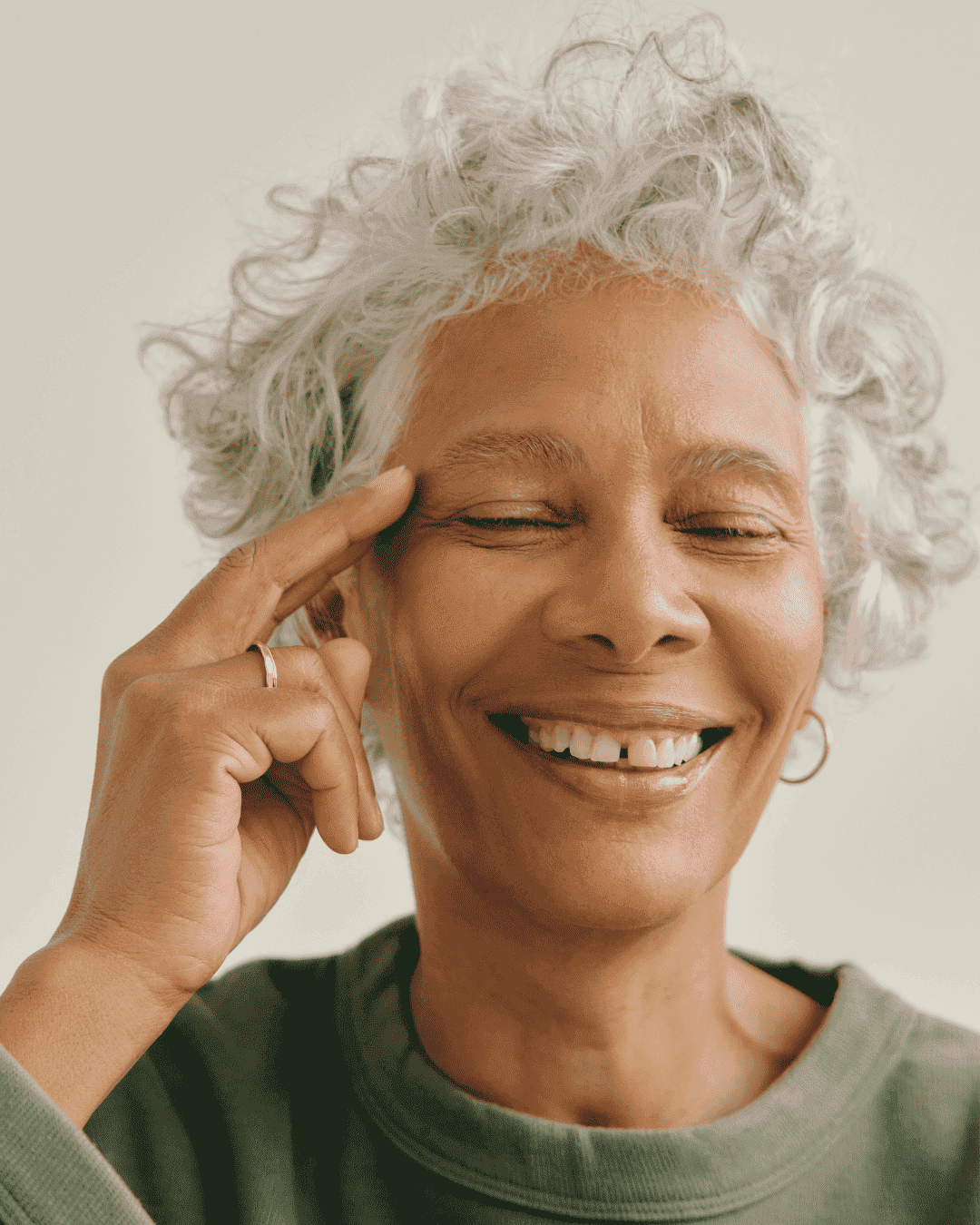 Eine dunkelhäutige Frau mit weißen Haaren und einem dunkelgrünen Pullover, die sich mit der rechten Hand auf die rechte Schläfe neben der Augenbraue greift. Sie hat die Augen dabei geschlossen und lacht.