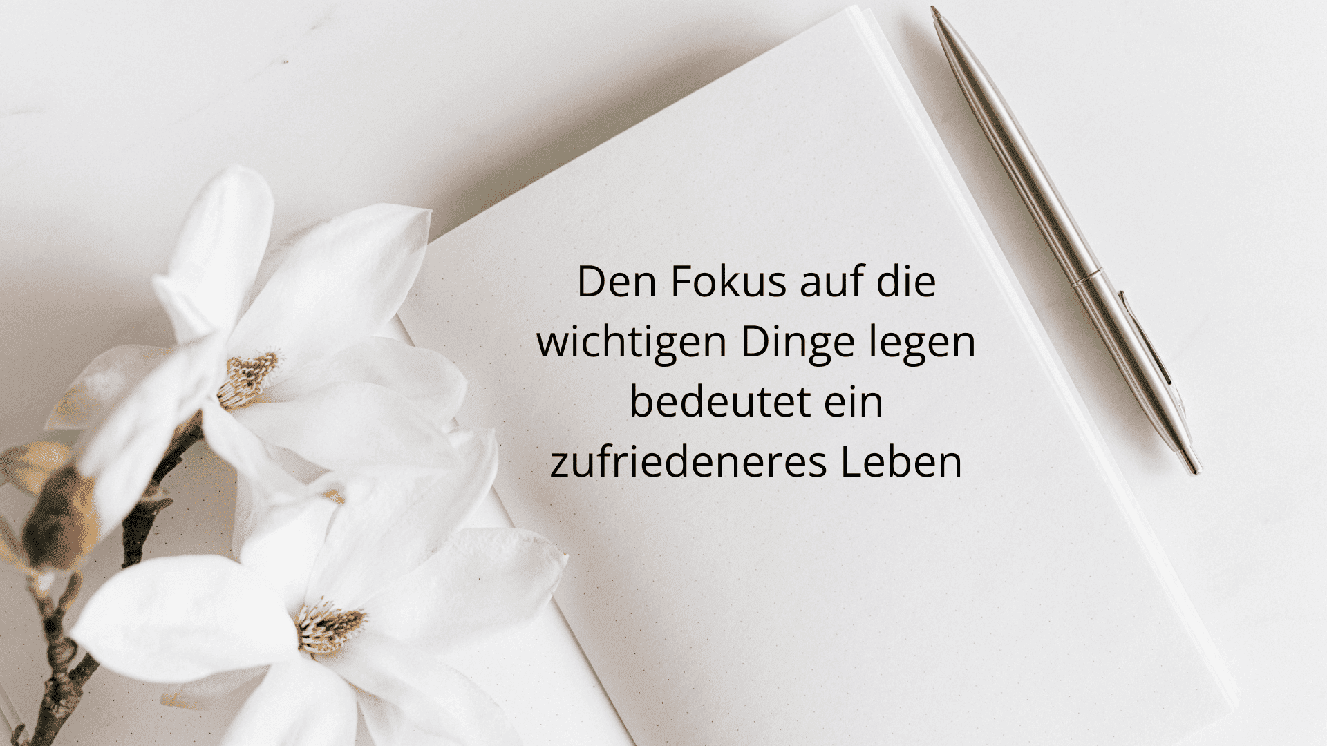 Den Fokus auf die wichtigen Dinge legen bedeutet ein zufriedeneres Leben. Auf einem weißen Tisch liegt ein Notizbuch, in dem dieser Text steht. Daneben sind weiße Orchideen, liegend.